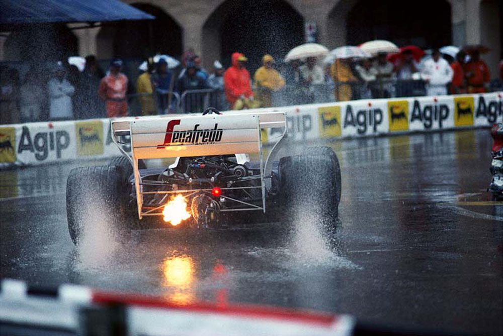 Formula 1 car emitting flames on a wet track during a race.