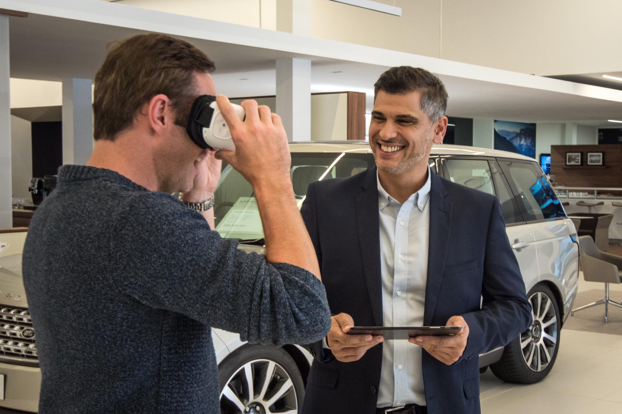 A man tries on VR goggles while another man in a suit smiles holding a tablet.