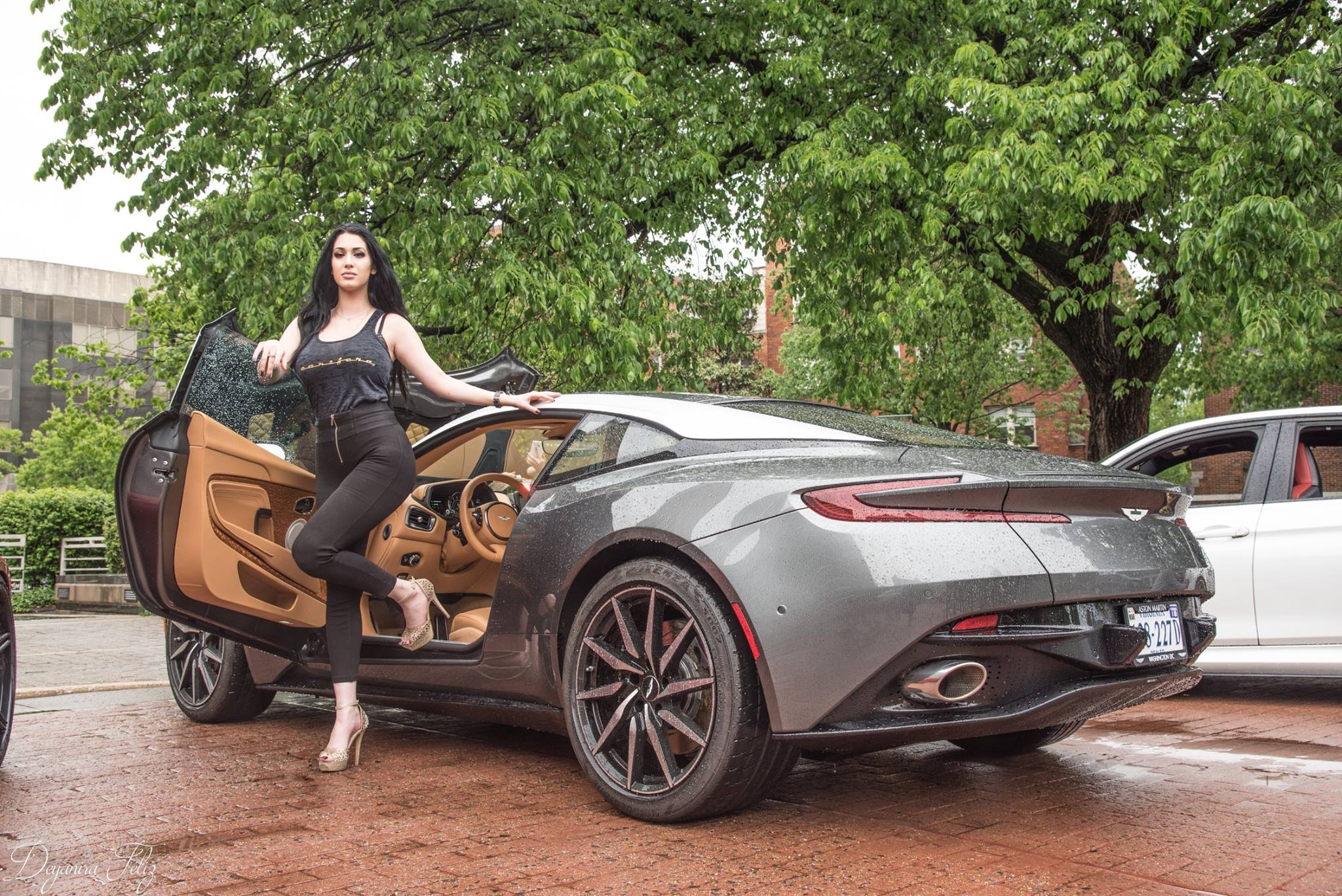 A woman in black poses by a sleek sports car under a leafy tree.