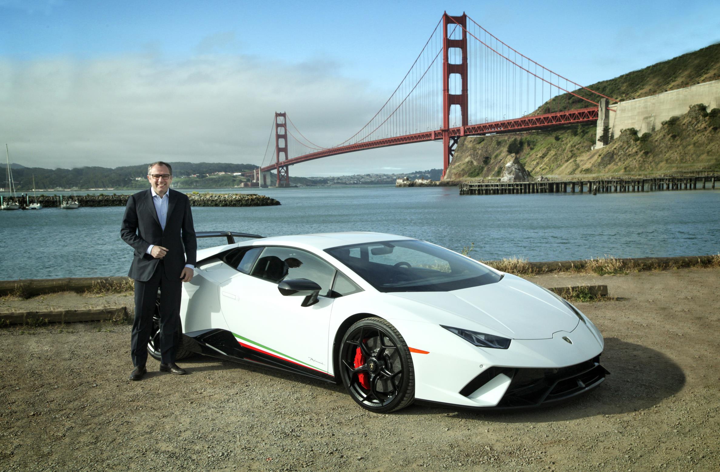 A man in a suit stands beside a white Lamborghini near the Golden Gate Bridge.