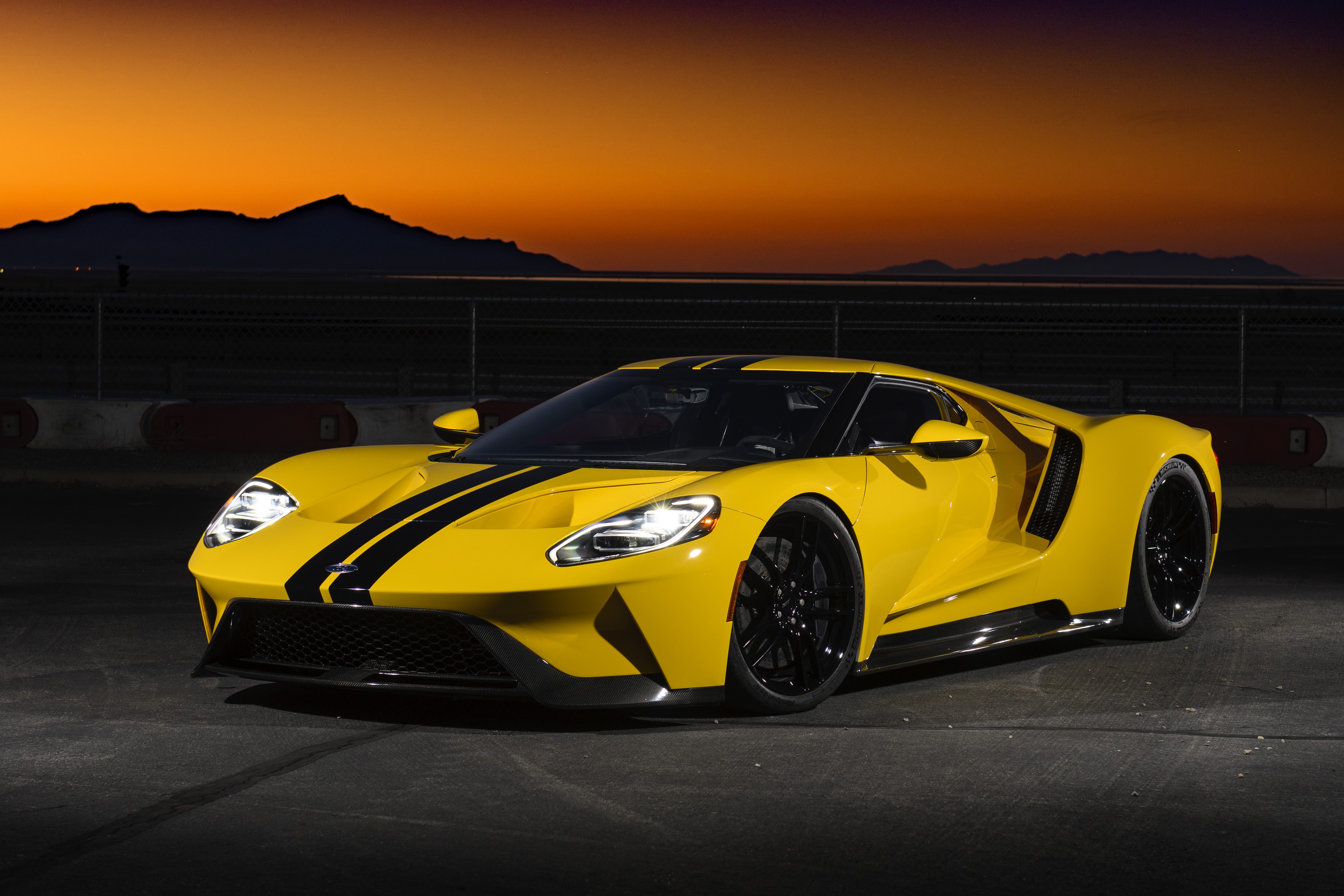 Yellow sports car with black racing stripes at sunset on a scenic road.
