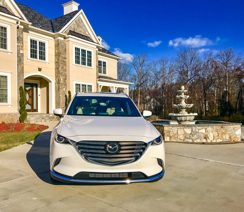 A white Mazda parked in front of a large house with a fountain.