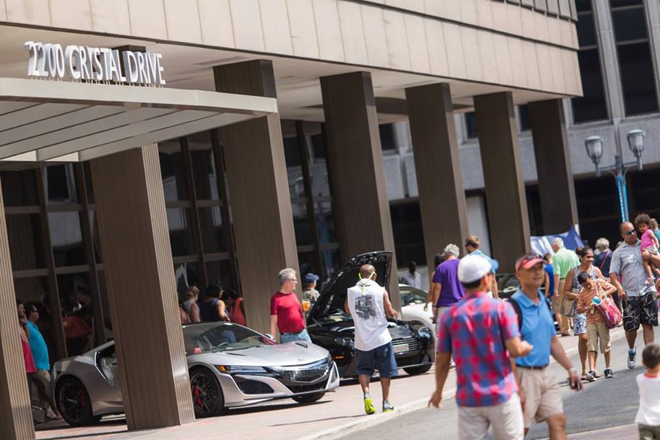 People gather outside a building entrance with a sports car parked nearby.