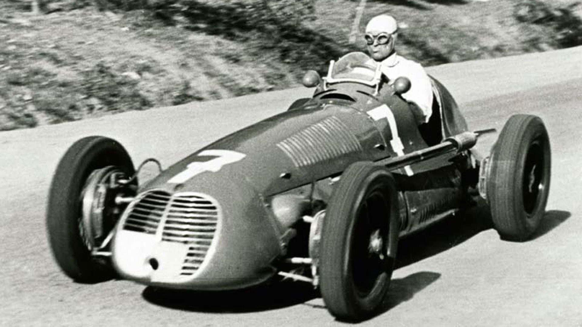 A vintage race car speeding on a road with a driver wearing goggles and helmet.