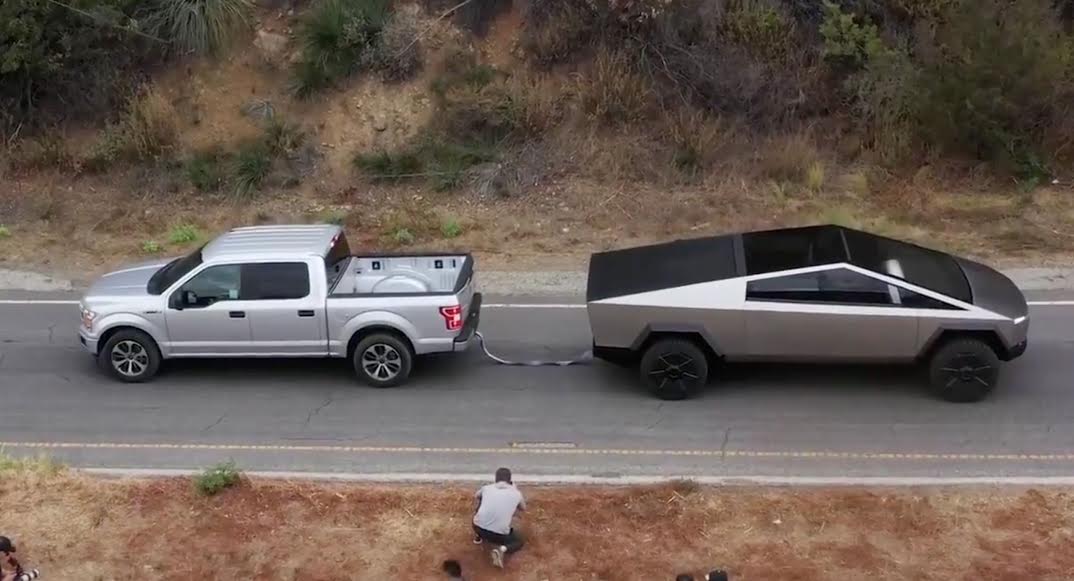 A person photographs a white pickup truck towing a futuristic black Tesla Cybertruck on a road.