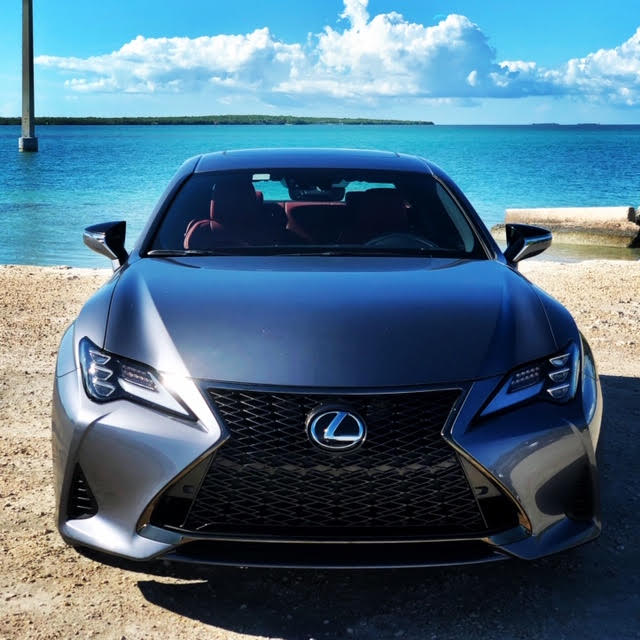 A sleek gray Lexus parked near a beach with clear blue water.