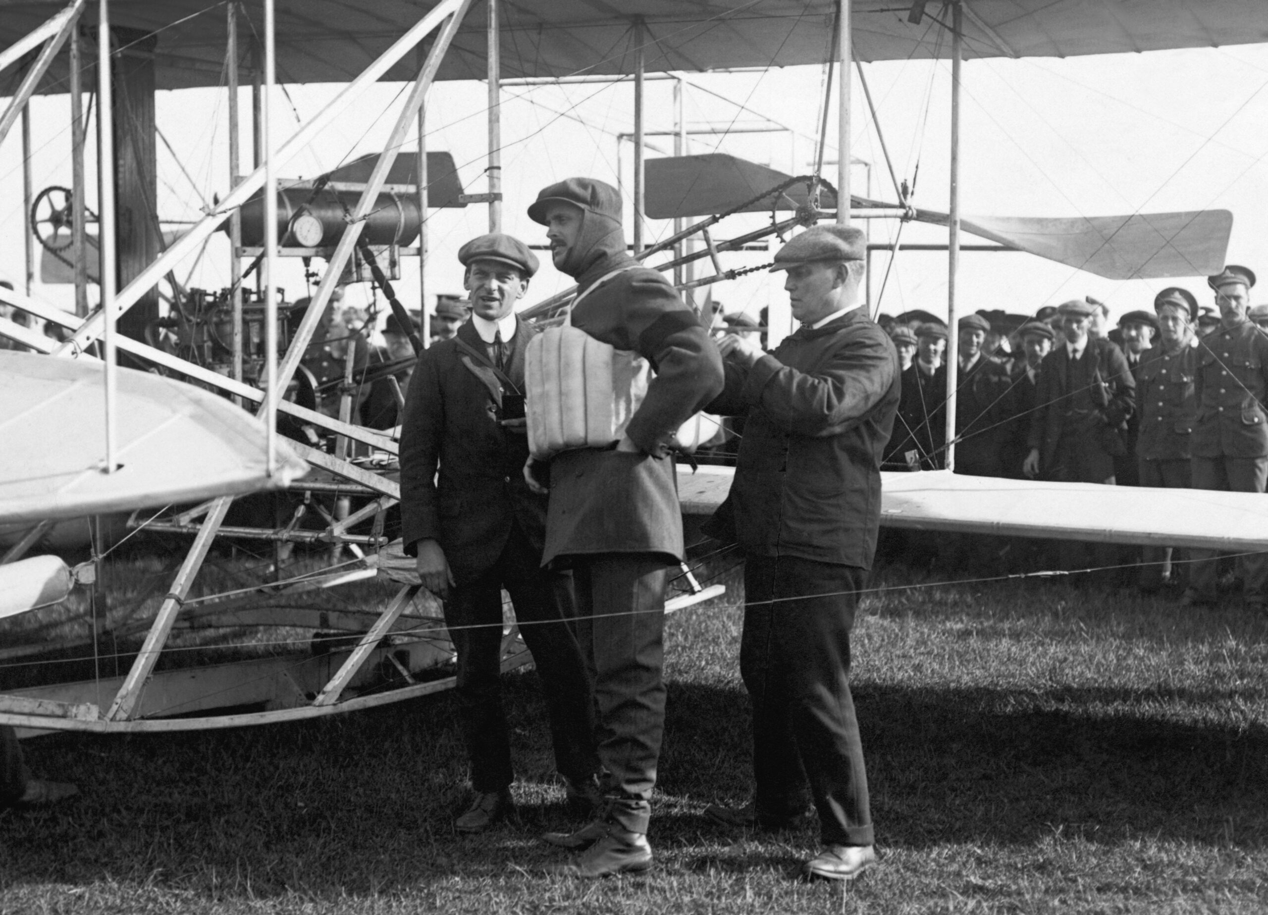 Three men in early 20th-century attire stand near a vintage biplane.
