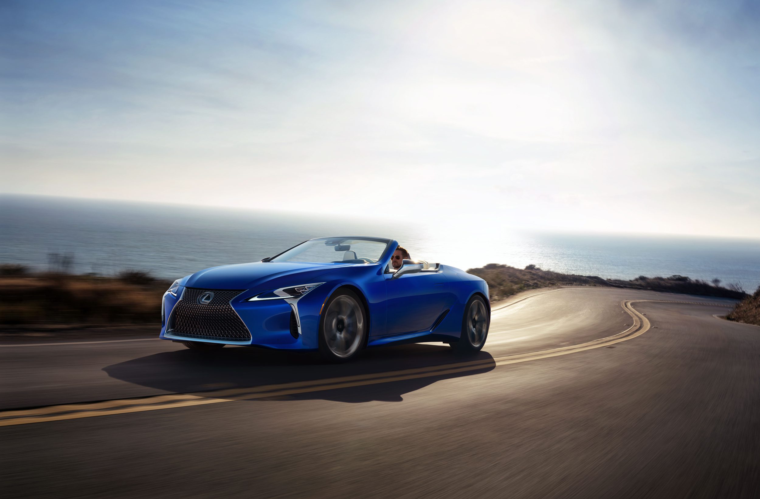 Blue convertible sports car driving on a coastal road during sunset.