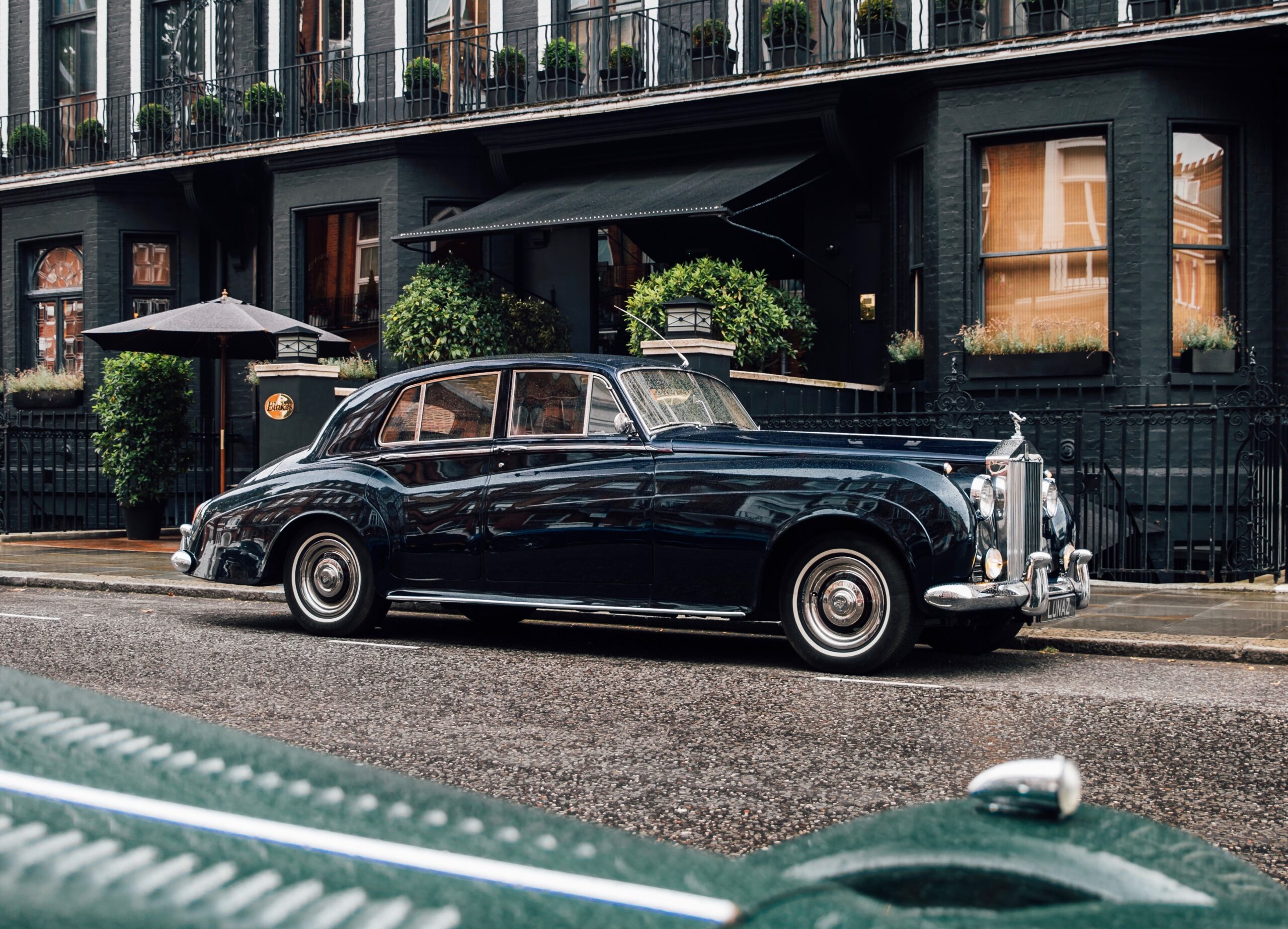 A classic black luxury car parked on a wet street beside a building with greenery.