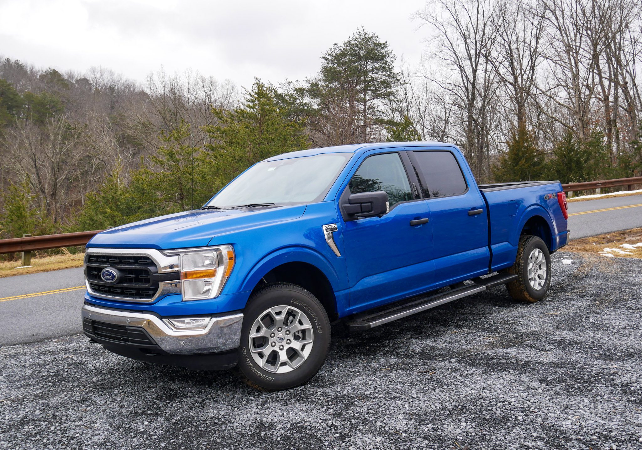 Blue Ford pickup truck parked on gravel with trees in background.