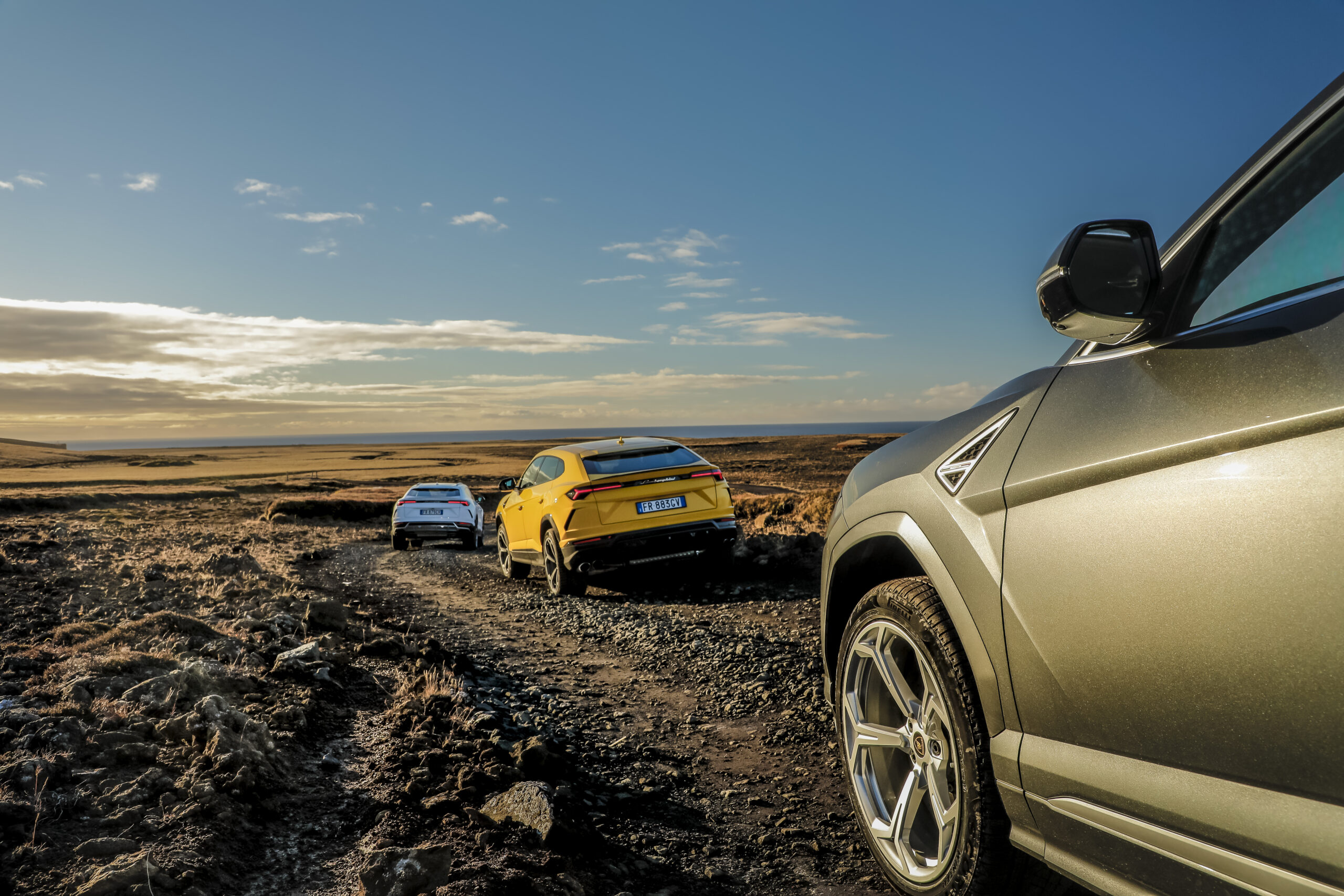 Three cars driving on a muddy off-road trail under a partly cloudy sky.