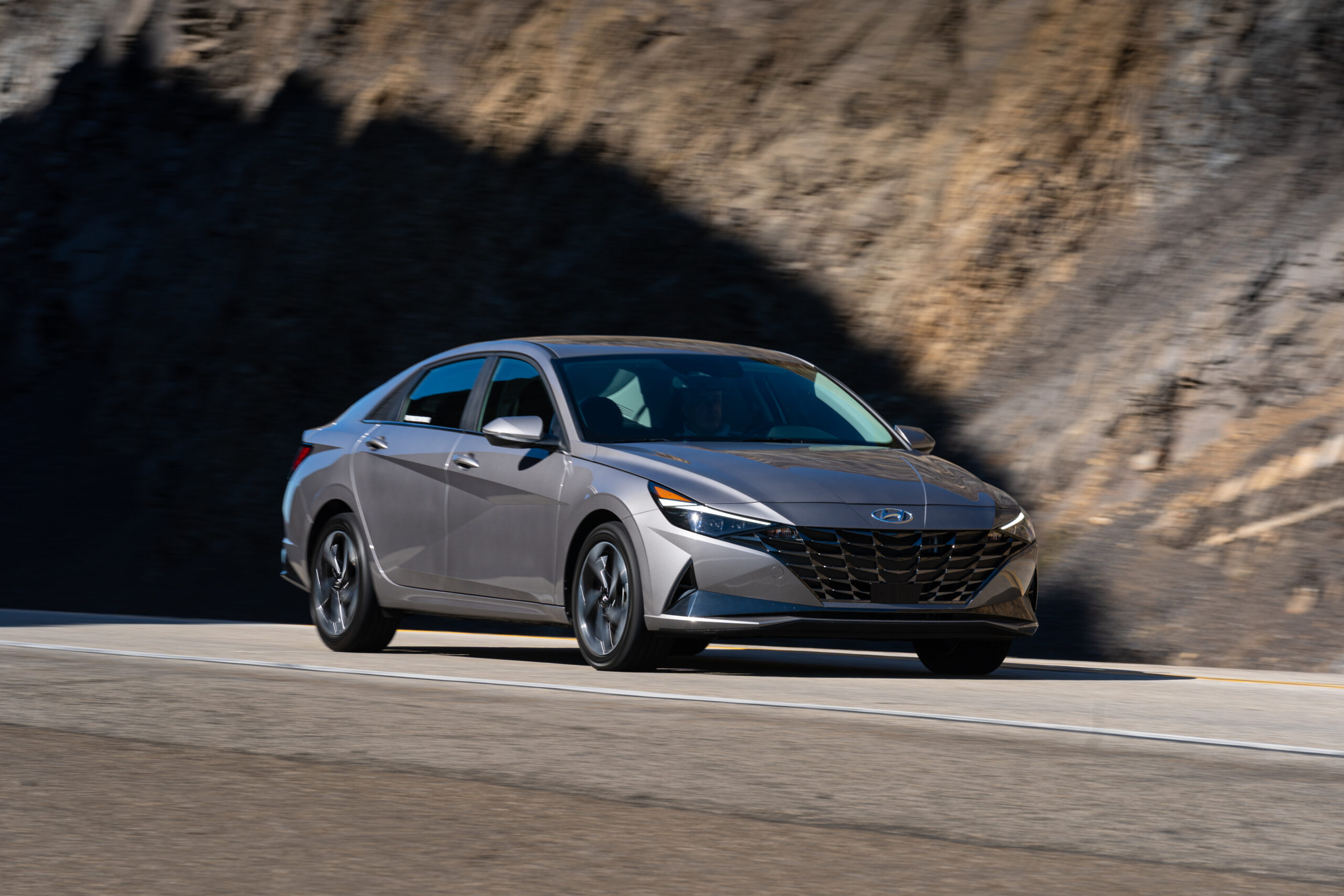 A sleek gray sedan driving on a winding road beside a rocky hill.