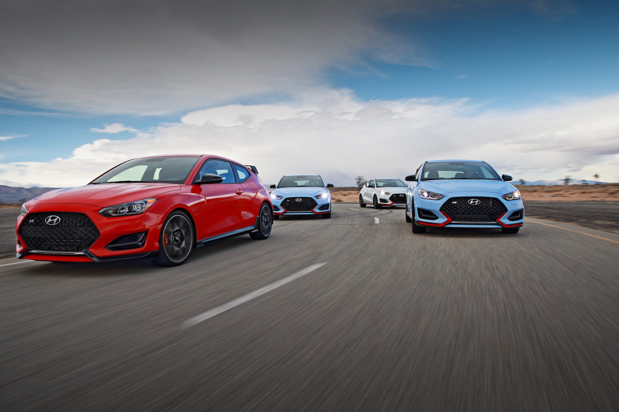 A lineup of sporty cars driving on a road under a cloudy sky.