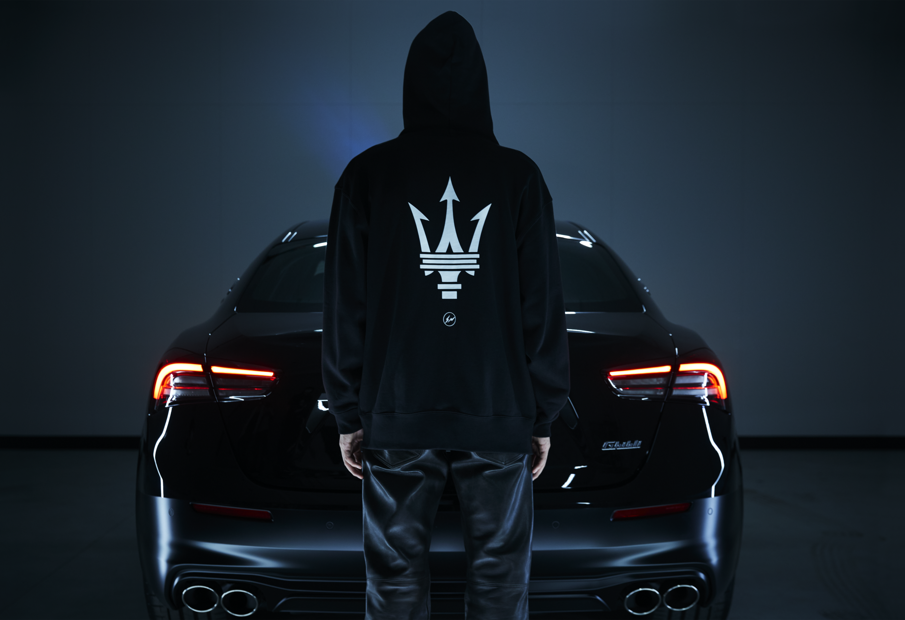 Person in black hoodie with trident logo standing before a luxury car at night.
