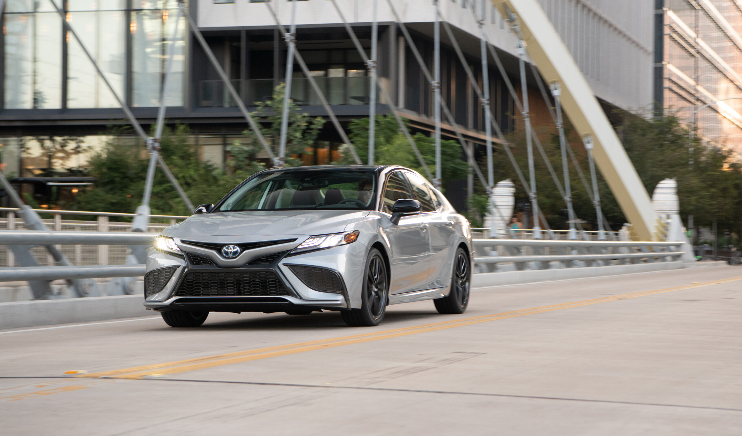 Silver Toyota Camry driving on an urban bridge with steel arches.