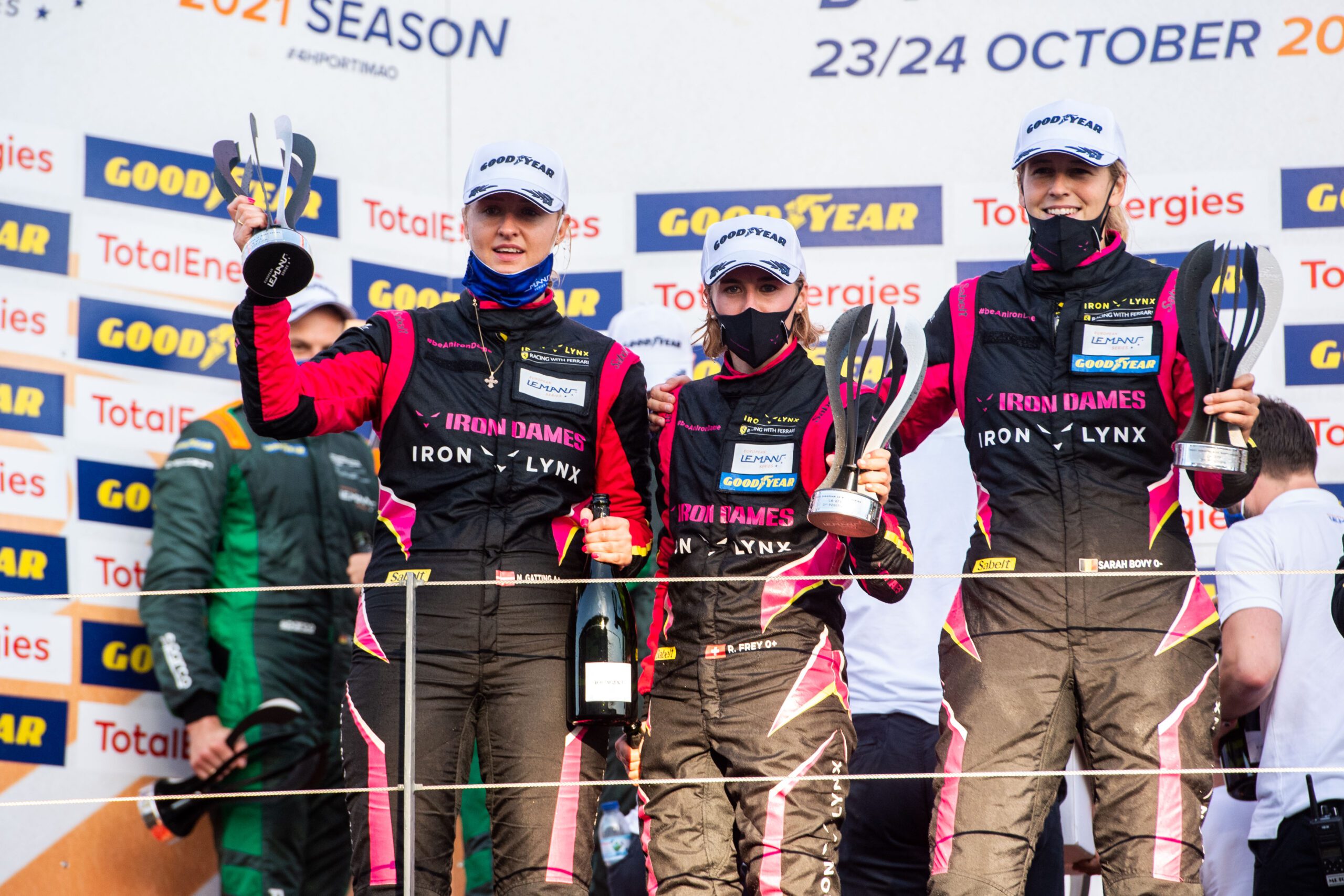 Four race car drivers celebrating on the podium holding trophies and champagne.