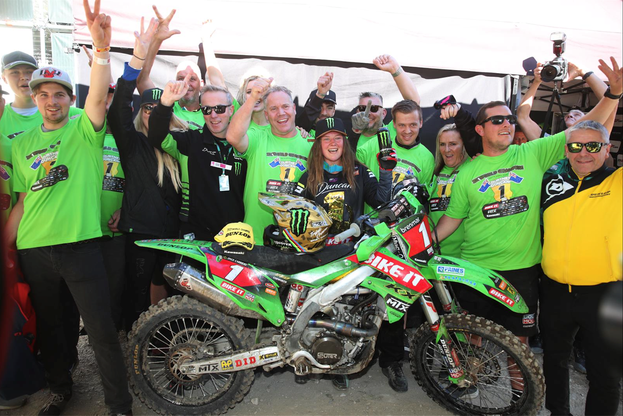 Motocross rider and team celebrate with raised hands around a green bike.