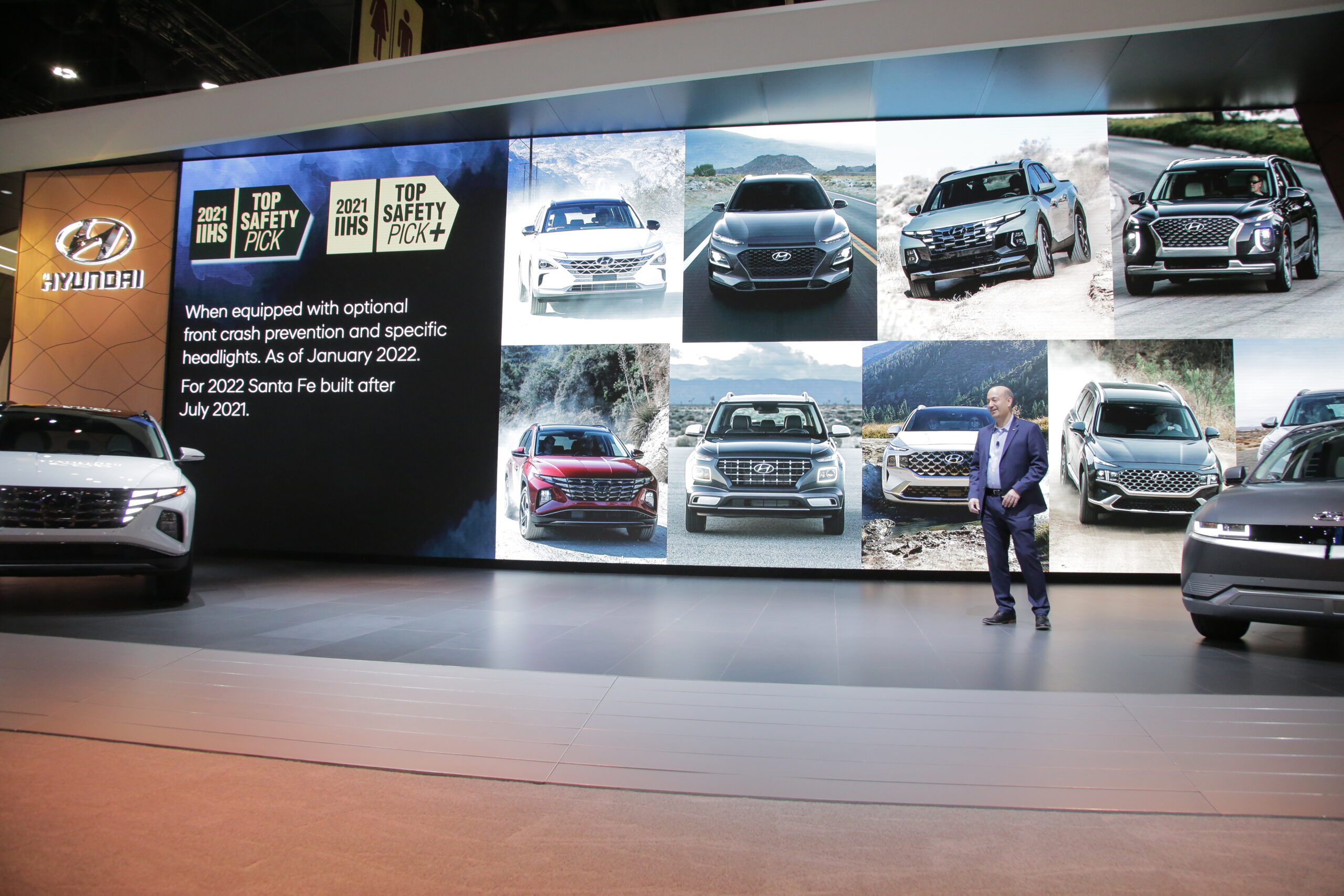 Presenter showcases various car models on a large screen at an auto event.