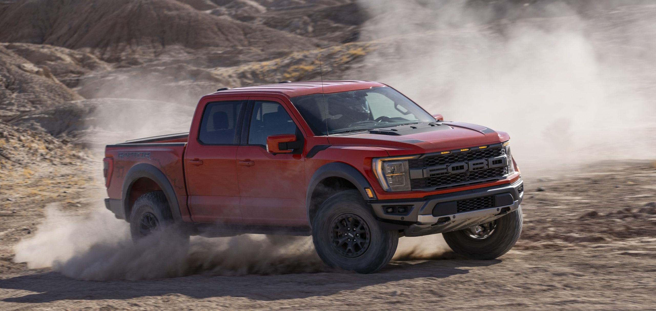 Red Ford Raptor pickup driving fast on a dusty desert road.