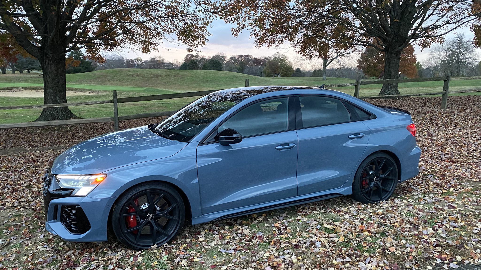 A sleek gray sports sedan parked on grass with a wooden fence and trees in the background.