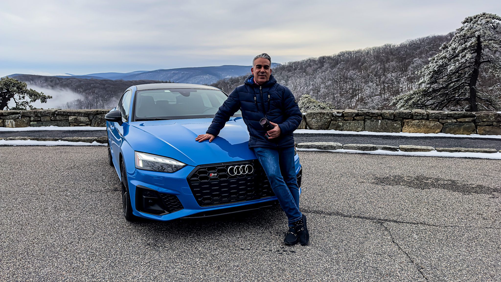 Man leaning on a blue Audi car on a mountain road.