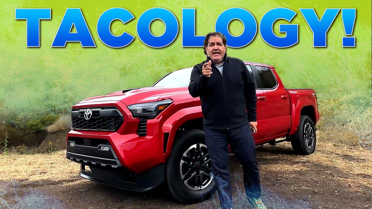 Man points while standing beside a red pickup truck in front of a colorful sign.