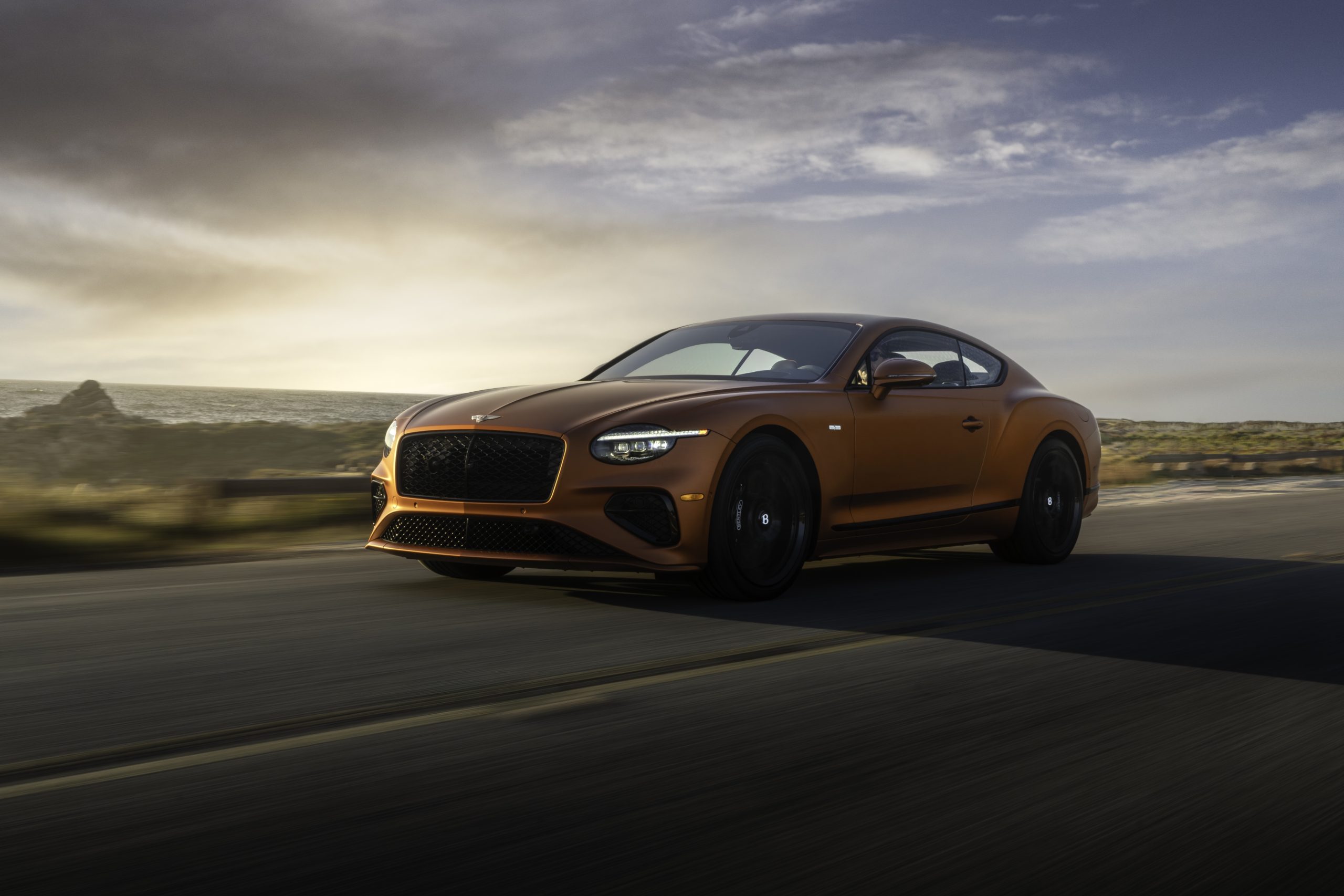A sleek, orange sports car driving on an open road under a cloudy sky.