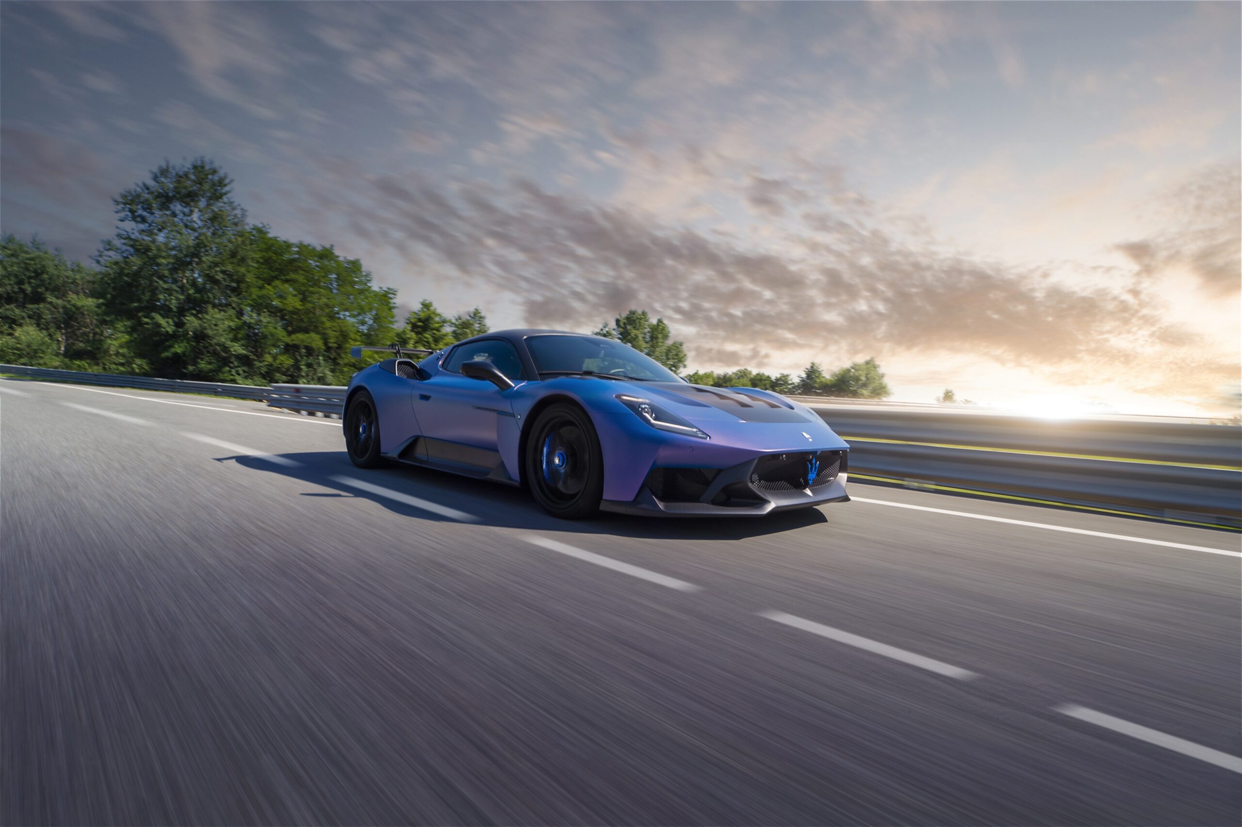 A blue sports car speeds along a road at sunset.