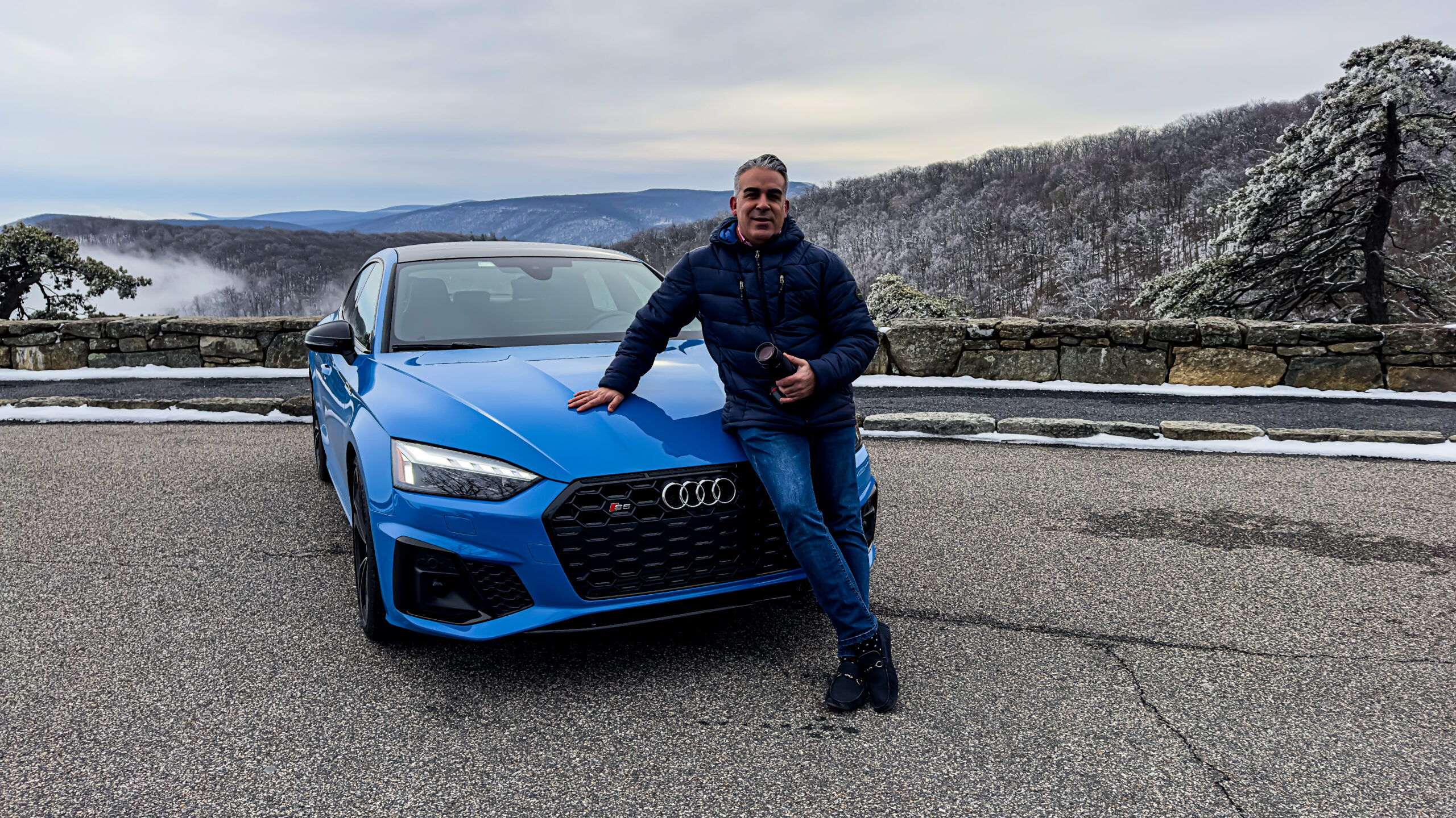 Man leaning against a blue Audi car on a scenic mountain road.
