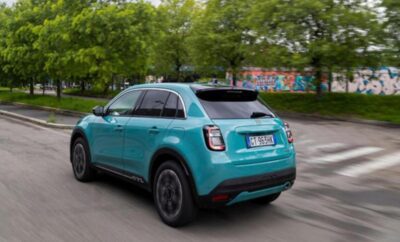 A teal compact SUV driving on a road with greenery and a graffiti wall in the background.