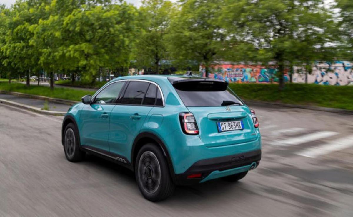 A teal compact SUV driving on a road with greenery and a graffiti wall in the background.