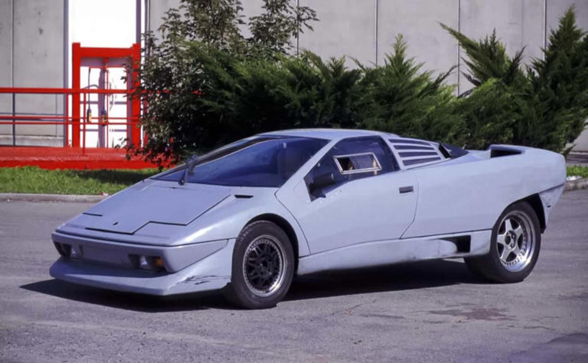 Futuristic silver car parked near bushes and a red structure.