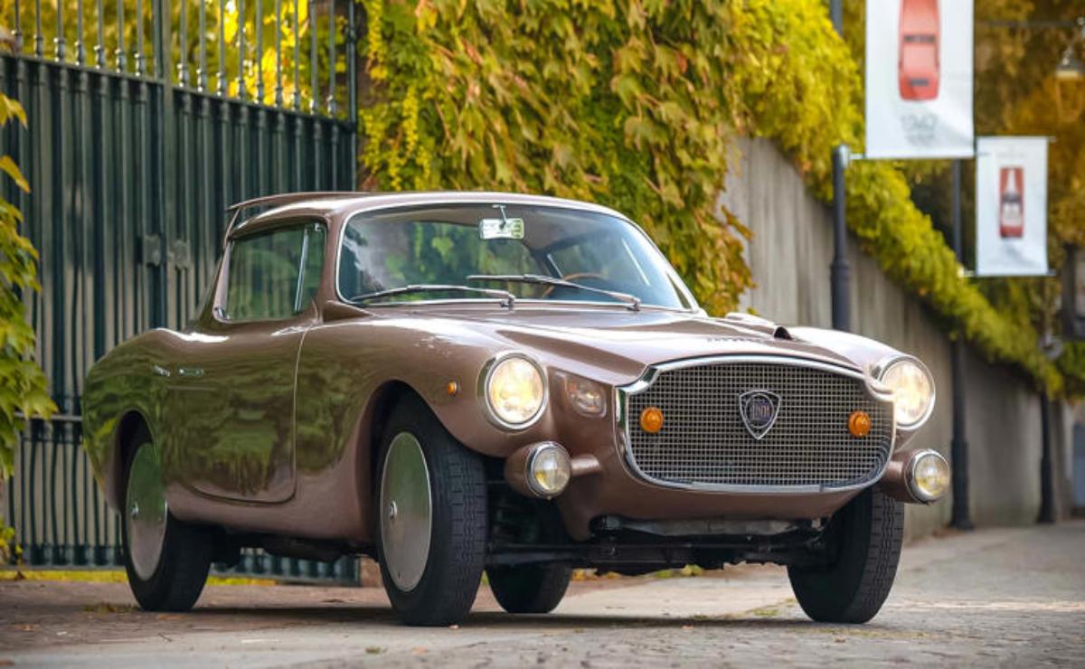 Vintage brown classic car parked near a green fence and yellow foliage.