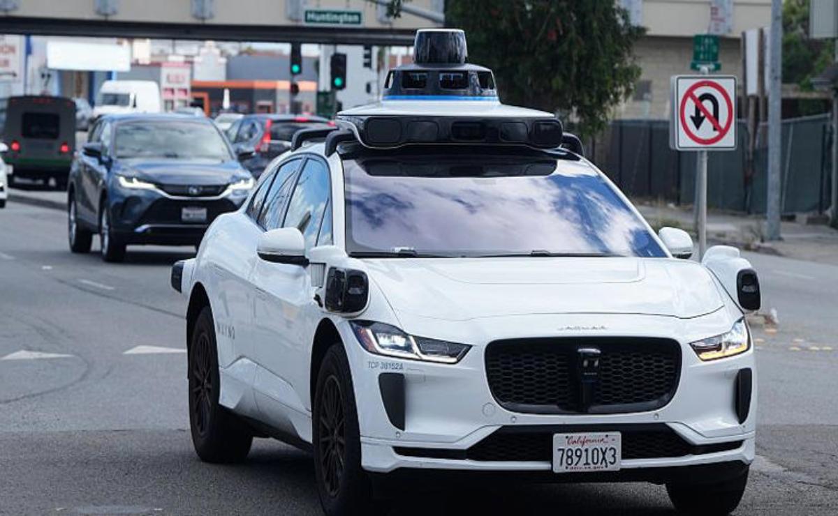 A white self-driving car equipped with rooftop sensors on a city street.