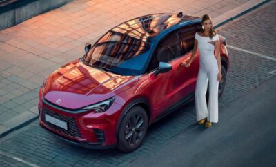 A woman in white stands next to a red electric SUV at sunset.