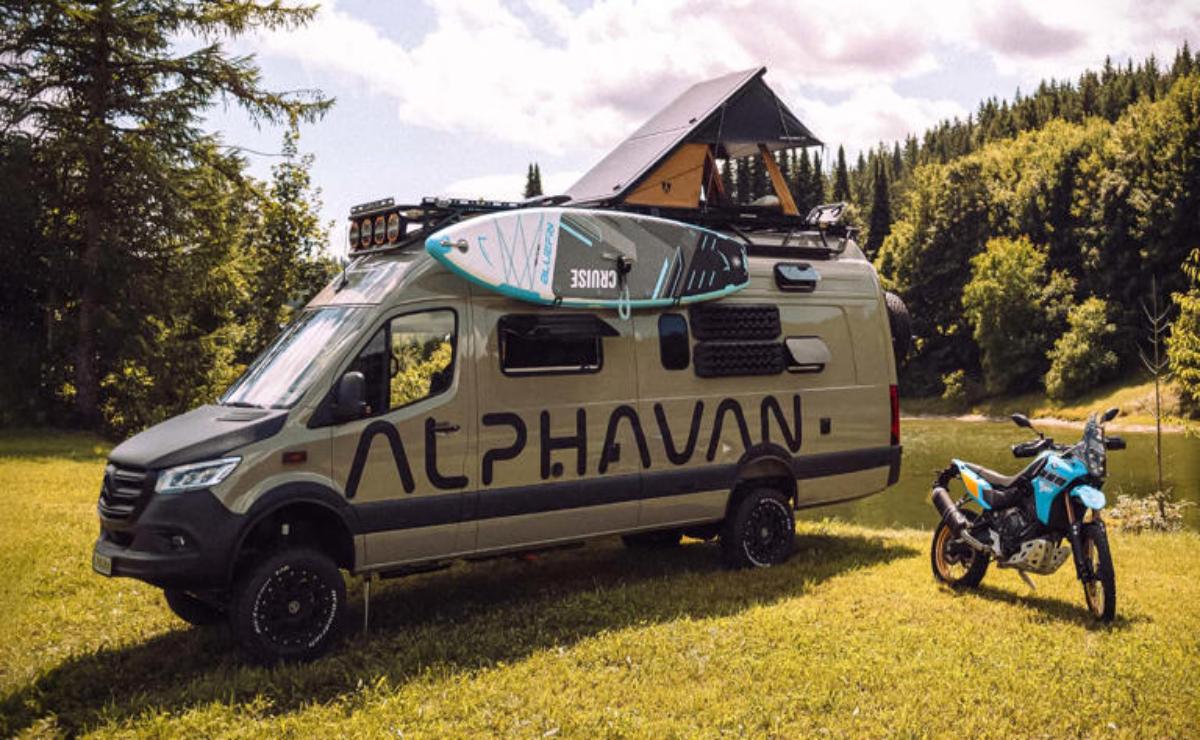 A camper van with a surfboard on top parked on grass near a wooden cabin.