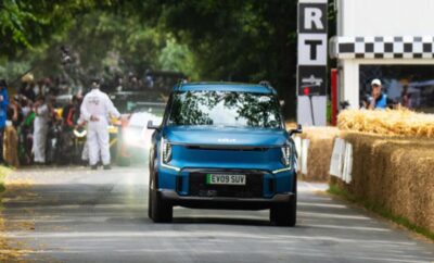 A blue electric SUV driving on a tree-lined road during a motorsport event.
