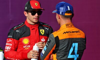 Two Formula 1 drivers in racing suits and caps chatting.