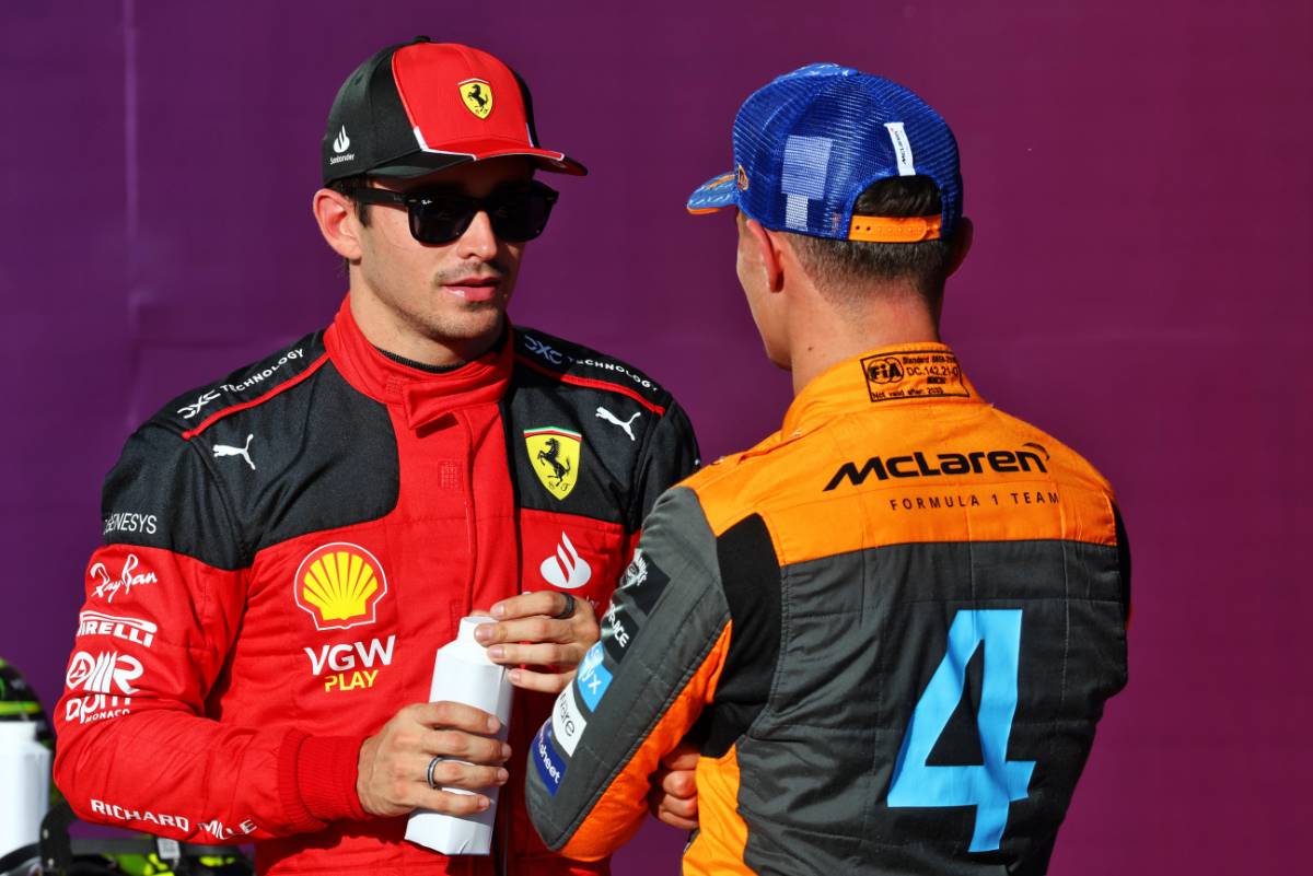 Two Formula 1 drivers in racing suits and caps chatting.