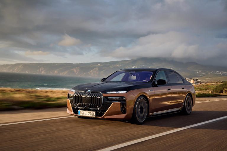 A sleek black luxury sedan driving on a coastal road under cloudy skies.