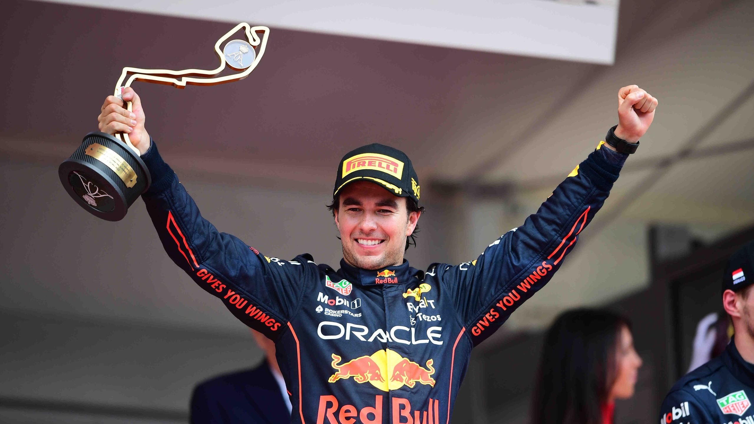 Race car driver celebrating victory on podium wearing Red Bull Oracle suit.