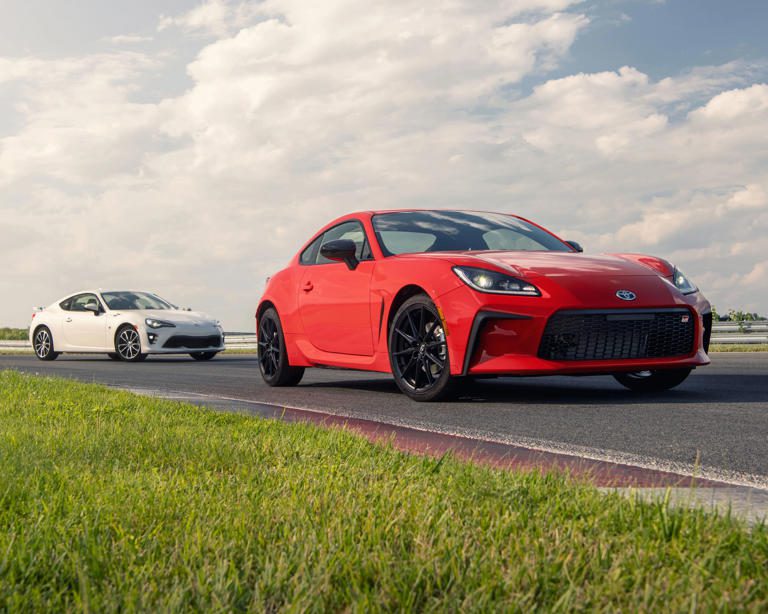 A red sports car races on a track with a white car behind it under a cloudy sky.