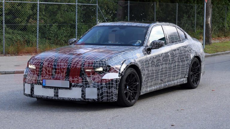 A camouflaged prototype car undergoing testing on a road.