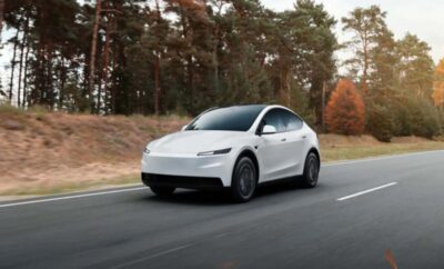 A white Tesla Model Y driving on a road surrounded by autumn trees.