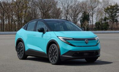 A modern turquoise electric car parked outdoors with leafless trees in the background.