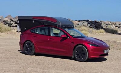 A sleek red Tesla Model Y parked on a sandy terrain with rocks in the background.