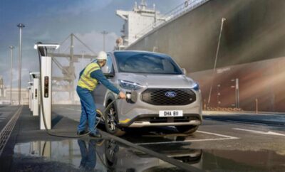 Worker cleaning a car at a port with ships in the background.