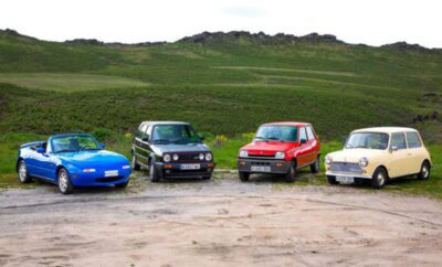 Four vintage cars parked on a dirt road with green hills in the background.