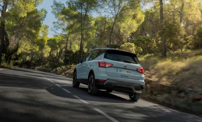 A light blue SUV driving on a winding forest road under sunny skies.