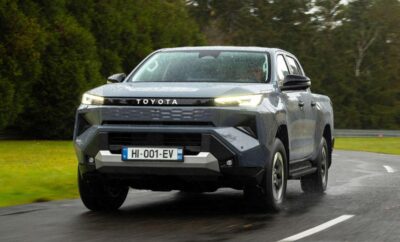 A silver Toyota pickup truck driving on a road with trees in the background.